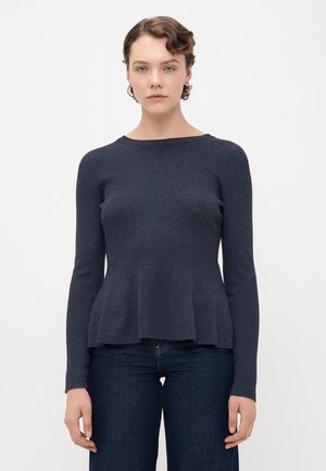 Woman with short wavy hair wearing a fitted, long-sleeve dark blue sweater and dark blue jeans, standing against a plain light background.