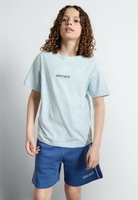 Camiseta de algodón de color azul claro con mangas cortas y cuello redondo, que presenta la palabra "elemento" en azul. Combinada con pantalones cortos de color azul marino.