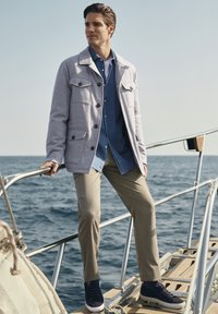 Helle graue Jacke mit großen Taschen, blaues Hemd mit Knöpfen, beige Hose und marineblaue Sneaker, steht auf einem Boot vor dem Ozean-Hintergrund.