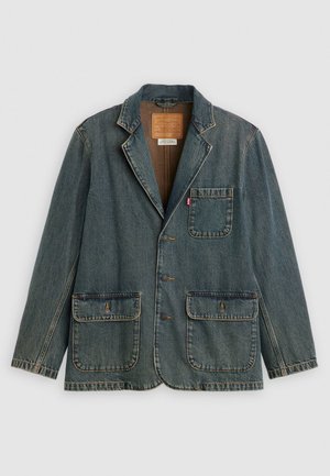 Blazer en denim avec trois boutons, une poche poitrine avec une étiquette rouge, et deux grandes poches avant avec rabat, posé à plat sur un fond blanc.