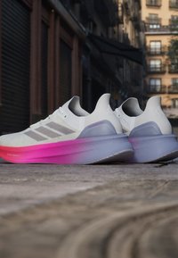 Scarpe sportive bianche con una suola che sfuma dal rosa al grigio. Presentano tre strisce grigie sul lato e una tomaia leggera e testurizzata.