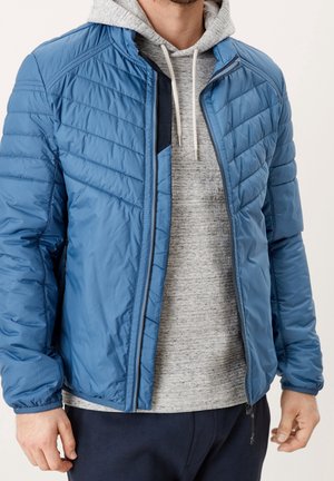 Uomo con giacca trapuntata blu aperta sopra una felpa con cappuccio grigio chiaro e pantaloni scuri, in piedi con le mani rilassate lungo i fianchi.