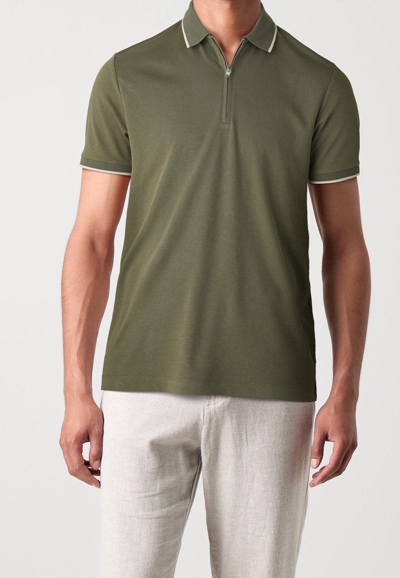 Polo-shirt vert olive avec une demi-fermeture éclair, col et poignets contrastés, fabriqué en tissu lisse. Porté avec un pantalon de couleur claire.