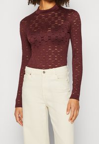 Kvinna iklädd en åtsittande långärmad bordeauxfärgad spetsblus med cirkulära mönster och högmidjade off-white byxor.