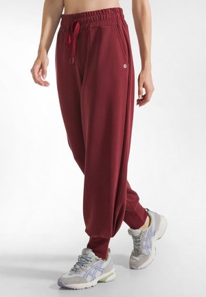Burgundy tapered joggers with elastic waistband and drawstring, featuring side pockets. Paired with light gray athletic sneakers.