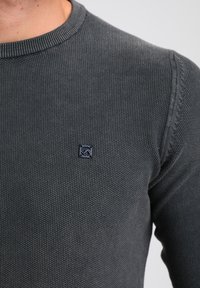 Pull gris foncé en maille avec un col rond côtelé et un petit logo bleu marine. Présente un tissu texturé avec des coutures visibles.