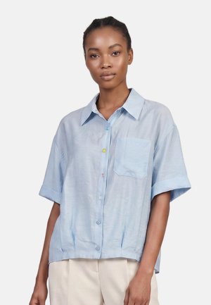 Giovane donna che indossa una camicia azzurro chiaro a maniche corte con bottoni e tasca frontale e pantaloni beige, in piedi contro uno sfondo bianco.