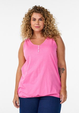 Kvinde med krøllet blondt hår iført en ærmeløs lyserød top og mørkeblå jeans, der viser en rosetatovering på venstre underarm.