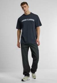 Tumši zils T-krekls ar "URBAN CLASSICS" logo, īsām piedurknēm, brīvs griezums, apvienots ar tumši pelēkiem džinsiem un gaišiem kedām.