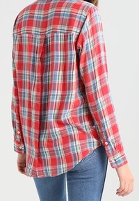 Camisa de cuadros roja y azul con mangas largas, puños abotonados y dobladillo redondeado. Fabricada con una tela ligera y con un detalle de pliegue en la parte posterior.