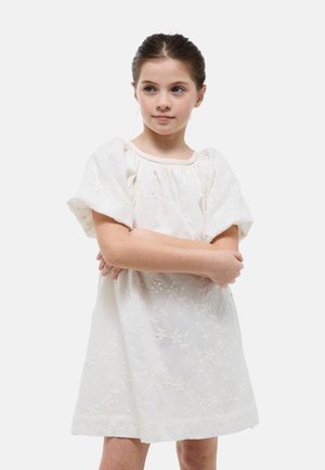 Fille aux cheveux bruns portant une robe blanche brodée de fleurs, debout les bras croisés, regardant légèrement sur le côté.