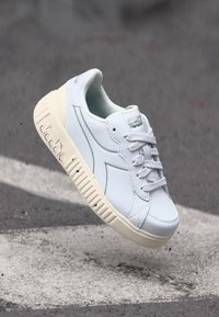 Fehér sneaker sima bőr felsőrésszel, lapos fűzőkkel és vastag krémszínű gumitalppal. Diszkrét márkázással és lekerekített orrformával rendelkezik.