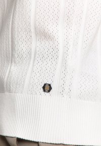 Witte gebreide trui met een gemêleerd patroon met kleine gaten, geribbelde zoom en een zwart hexagonaal logo met gouden strepen.
