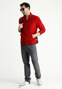 Sudadera de felpa roja con cremallera y bolsillo frontal, combinada con jeans grises y zapatillas blancas. El modelo lleva gafas de sol negras y lleva una mochila.