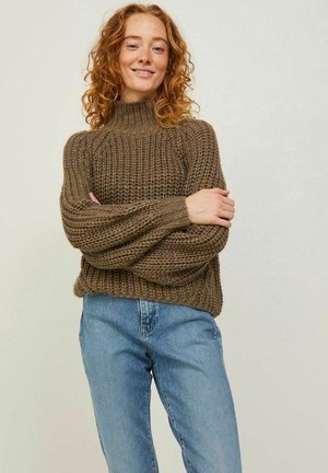 Brun striktrøje med høj hals, struktureret mønster, ballonærmer og afslappet pasform, parret med blå jeans.