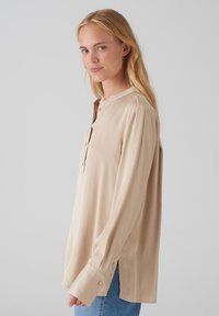 Beige Bluse mit langen Ärmeln, rundem Kragen, Knopfleiste und seitlichen Schlitzen. Glatte Textur, leichtes Material und goldfarbene Knopfdetails.