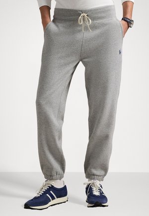 Man wearing gray sweatpants with elastic cuffs and drawstring, paired with white socks and blue athletic shoes, hands in pockets.