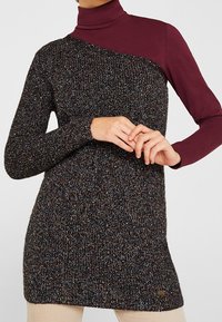Zwarte en veelkleurige gebreide sweater jurk met een col. Het heeft een asymmetrisch ontwerp met een effen bordeauxrode sectie op één schouder.
