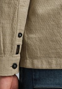 Langarmshirt in beige textuurstof. Het heeft zwarte knopen en een branded label nabij de zoom. Gekleed met donkere denim zichtbaar hieronder.
