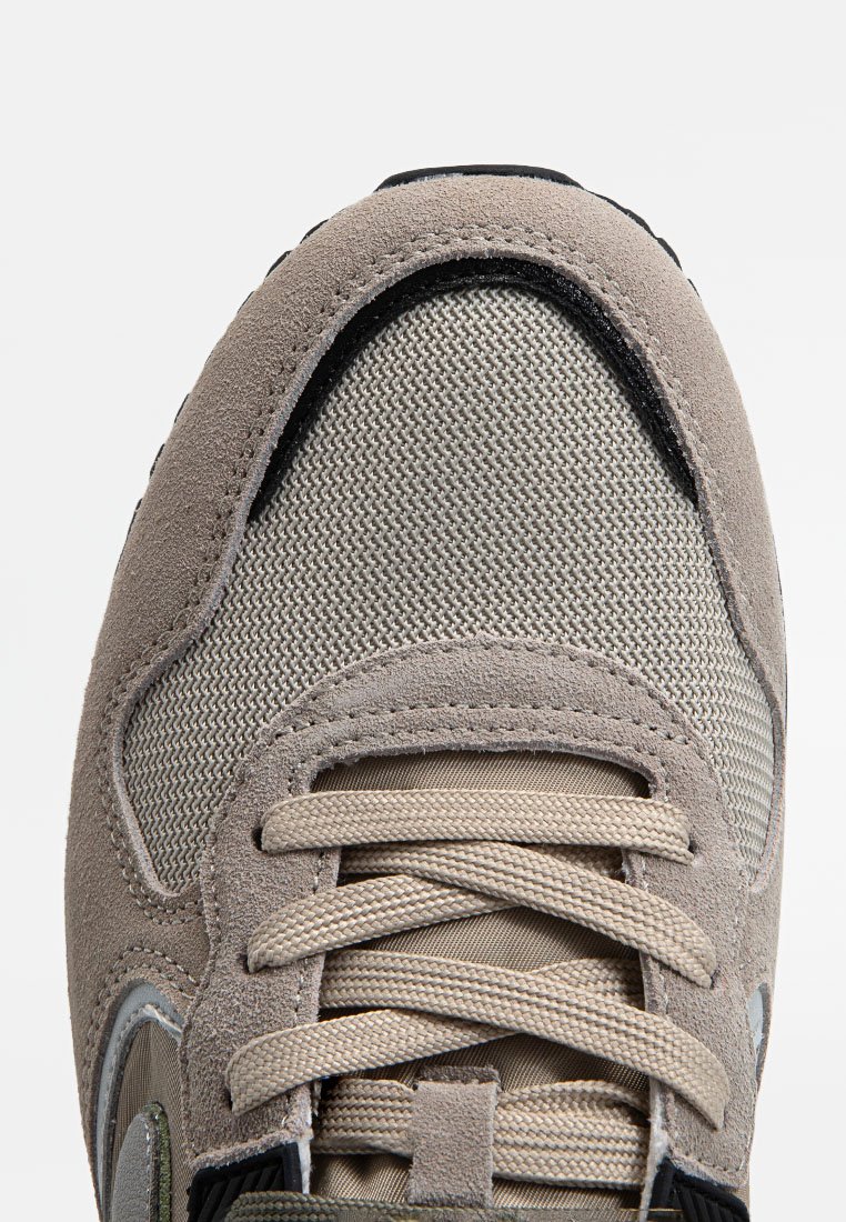 Grijze sneaker met suède en mesh materialen, ronde veters in beige, een gestructureerd oppervlak en een zwarte accentrand langs de punt.