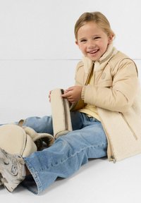 Lachend kind in beige schapenvachtjack en spijkerbroek zit op de vloer en houdt een bijpassend beige etuitje met rits vast, naast zich staat een rugzak.