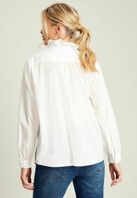 Weißes Langarmhemd mit Rüschenausschnitt, gerafftem Rücken und geradem Saum, getragen mit dunkelblauen Jeans. Glattes, leichtes Material.