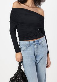 Black off-the-shoulder top with ruched texture and long sleeves, paired with light blue high-waisted jeans. Holding a black floral bag.