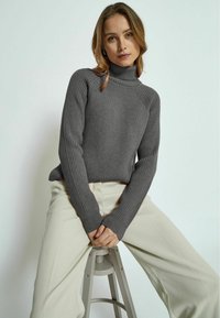 Gray ribbed turtleneck sweater with long sleeves, paired with light beige wide-leg pants, seated on a gray stool against a light background.