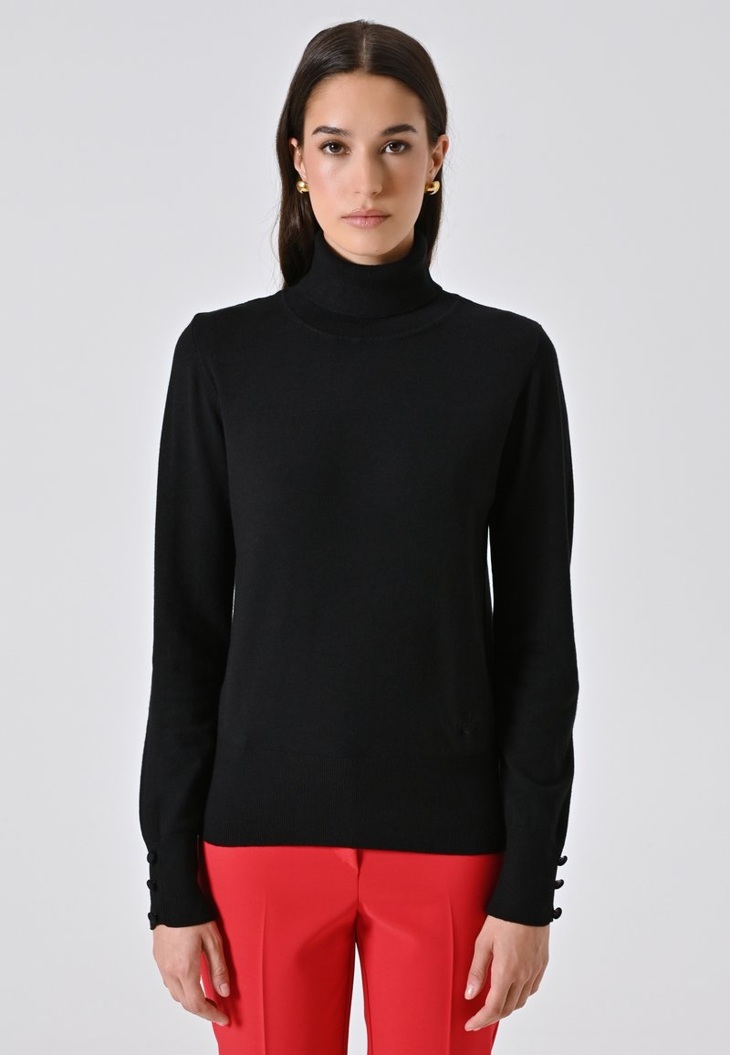 Frau mit einem schwarzen Rollkragenpullover mit Knopfdetails an den Ärmeln und leuchtend roten Hosen vor einem schlichten hellen Hintergrund.