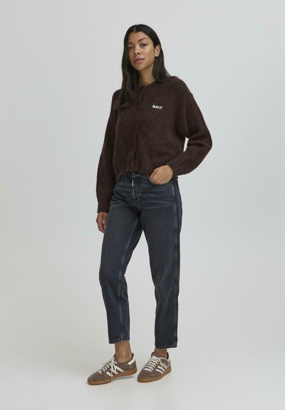 Brązowy sweter dzianinowy z guzikami, z małym białym logo. Noszony z ciemnymi, wysokimi jeansami oraz brązowymi sneakersami. Prosta faktura.