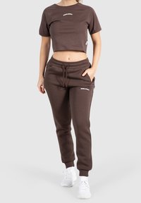 Frau trägt ein braunes, kurzes T-Shirt und passende Jogginghose mit weißen Sneakers, steht vor einem einfarbigen, hellen Hintergrund.