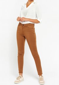 Slim-fit brown pants made of soft cotton blend, featuring front pockets, and paired with a white collared shirt and beige sneakers.