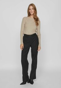 Pulover tricotat cu model în relief de culoare bej, asortat cu pantaloni negri, bine croiți, cu o textură netedă. Pantofii negri cu vârful ascuțit completează ținuta.
