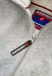 Grauer Sweatshirt mit Reißverschluss und weichem, strukturiertem Innenfutter, ausgestattet mit einem silbernen Reißverschluss und einem schwarzen Etikett mit "SUPERDRY" in Weiß.