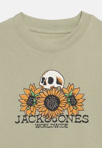 Ανοιχτό πράσινο μπλουζάκι με γραφικό κρανίο πίσω από τρία πορτοκαλί ηλιοτρόπια και κείμενο «JACK & JONES WORLDWIDE» από κάτω.