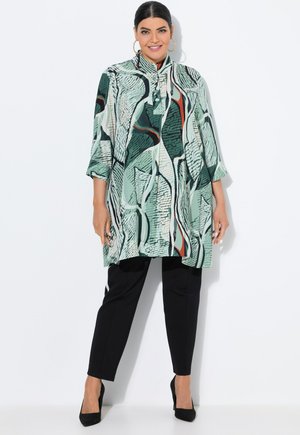 Femme portant un pantalon noir et un tunique à manches longues à motif abstrait vert avec un col haut et des talons noirs, souriant sur un fond blanc.