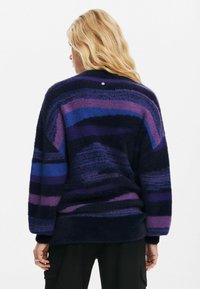 Getextureerde oversized trui in marineblauw, paars en blauwe strepen. Heeft een ronde halslijn en een klein hartdetail op de achterkant.