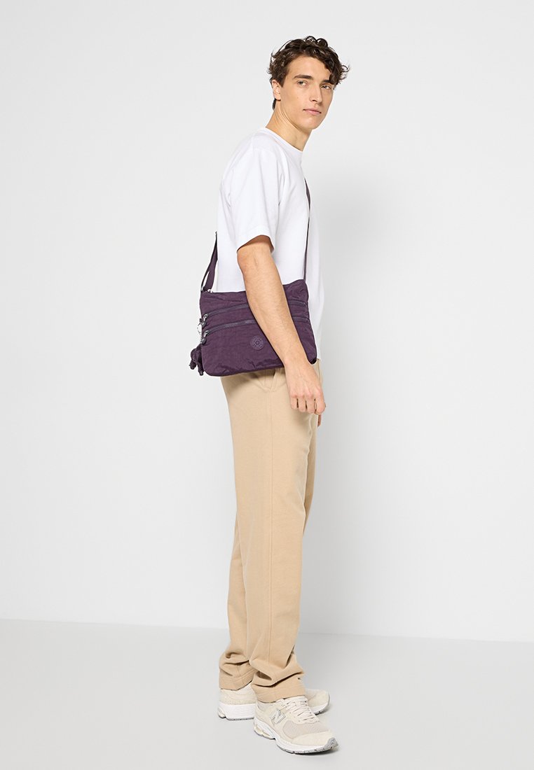 Borsa a tracolla viola realizzata in tessuto resistente, con multiple tasche con zip, una tracolla regolabile e un dettaglio con logo circolare.