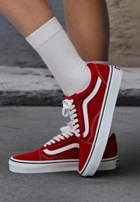 Baskets montantes en daim rouge avec accents blancs, bordures noires et lacets blancs. Portées avec une chaussette blanche à la cheville, montrant un matériau texturé et des lignes de design.