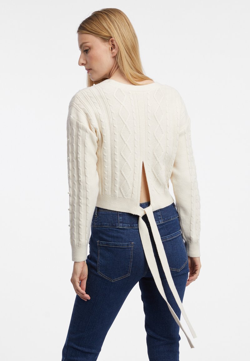 Pull en tricot crème avec un design en cable texturé, dos ouvert avec une fermeture à nouer, et manches longues avec des boutons d'accent.