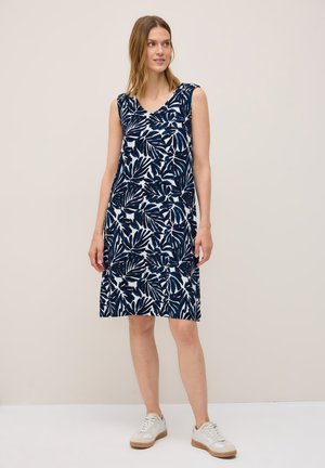 Model wearing a sleeveless navy dress with a white leaf pattern, paired with white sneakers, standing against a neutral background.