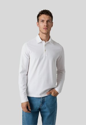 Jonge man die een witte lange mouwen polo en blauwe spijkerbroek draagt, staande tegen een effen lichtgrijze achtergrond met één hand in zijn broekzak.
