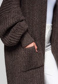 Ruskea neulecardigan, jossa on teksturoidut resorit, ylisuuret hihat ja sivutaskut. Kangas näyttää pehmeältä ja lämpimältä.