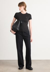 T-shirt noir ajusté à manches courtes, associé à un pantalon noir à jambes larges avec un léger scintillement. Le modèle tient une grande pochette noire.