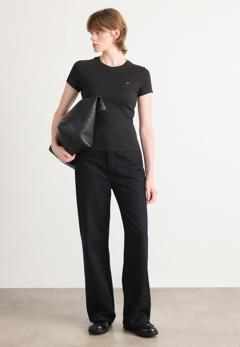 T-shirt noir ajusté à manches courtes, associé à un pantalon noir à jambes larges avec un léger scintillement. Le modèle tient une grande pochette noire.