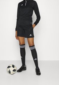 Conjunto deportivo negro con camiseta de manga larga y cremallera y pantalones cortos, combinado con calcetines negros hasta la rodilla con rayas blancas y botas de fútbol negras.