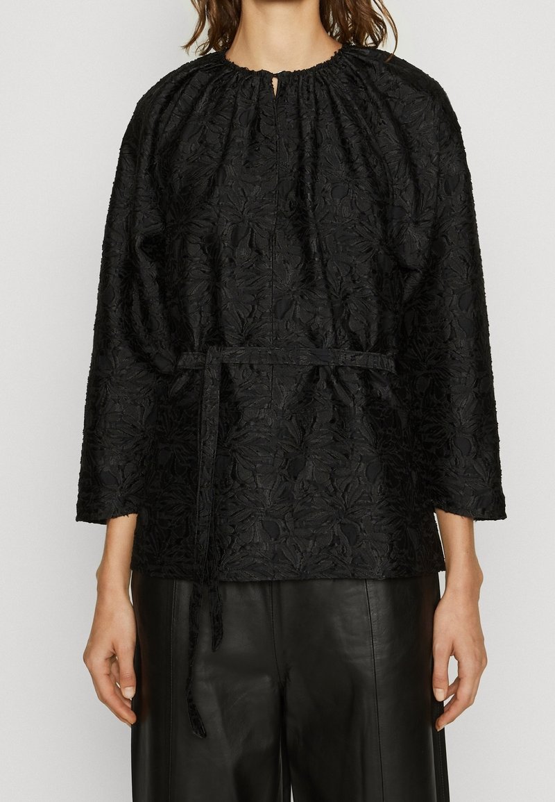 Blouse noire texturée avec motif floral, encolure froncée et ceinture à nouer. Comprend des manches longues et une ouverture partielle à l'avant.