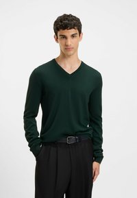 Grüner V-Ausschnitt-Pullover aus feinem Strickmaterial, mit langen Ärmeln und schlanker Passform, kombiniert mit schwarzen schmalen Hosen und einem Gürtel.
