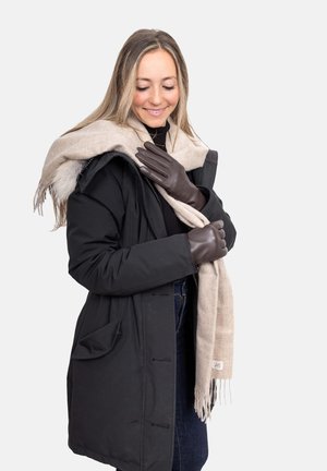 Schwarzer Parka mit sichtbaren Taschen, hellbeiger Schal mit Fransen und dunkelbraune Lederhandschuhe vor neutralem Hintergrund.