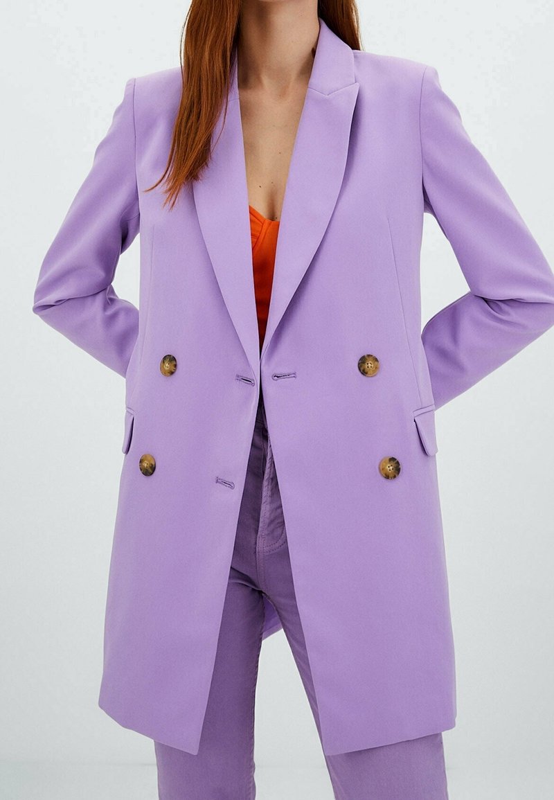 Femme portant un blazer lavande à double boutonnage avec des boutons marron sur un haut orange et un pantalon lavande assorti, les mains derrière le dos.